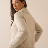 A woman wearing a cream-colored ribbed knit cardigan sweater, shown from a three-quarter back view against a solid beige studio background.