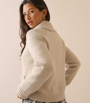 A woman wearing a cream-colored ribbed knit cardigan sweater, shown from a three-quarter back view against a solid beige studio background.