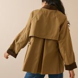 A woman shown from the back wearing a tan trench coat with dark brown contrast trim on the collar, cuffs, and pockets, paired with blue jeans against a solid beige studio background.