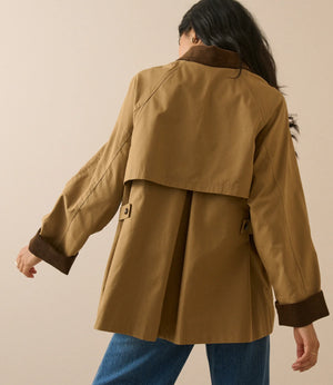 A woman shown from the back wearing a tan trench coat with dark brown contrast trim on the collar, cuffs, and pockets, paired with blue jeans against a solid beige studio background.