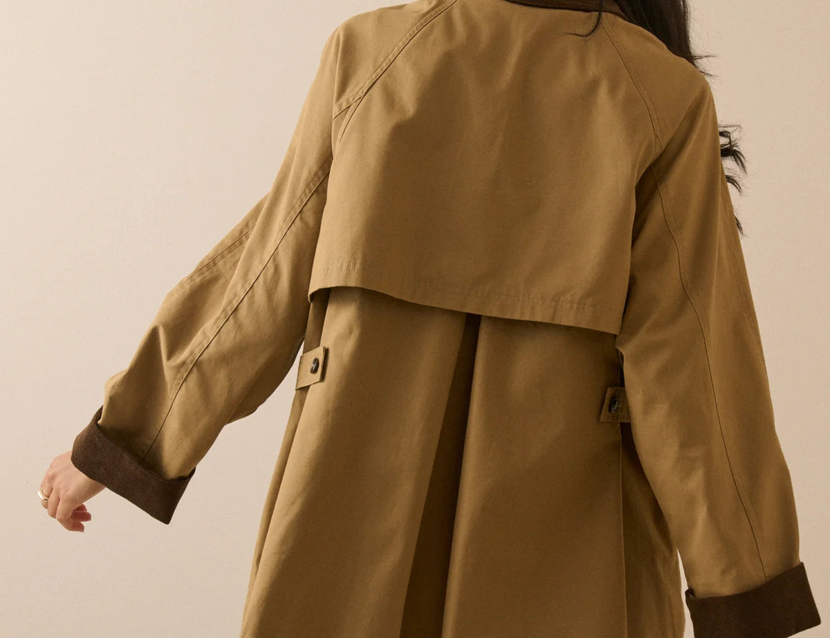 A woman shown from the back wearing a tan trench coat with dark brown contrast trim on the collar, cuffs, and pockets, paired with blue jeans against a solid beige studio background.