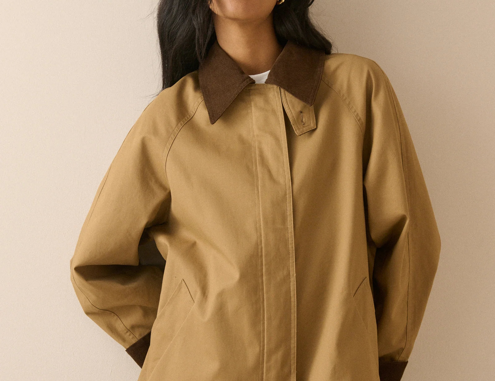A woman wearing a tan oversized utility jacket with contrasting dark brown collar, cuffs, and hem, paired with blue jeans, set against a plain beige background.