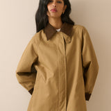 A woman wearing a tan oversized utility jacket with contrasting dark brown collar, cuffs, and hem, paired with blue jeans, set against a plain beige background.