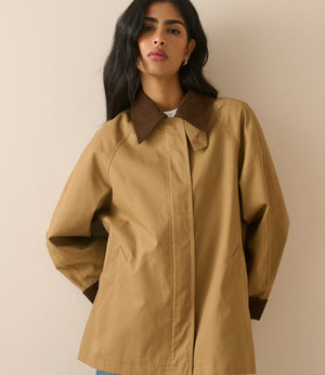 A woman wearing a tan oversized utility jacket with contrasting dark brown collar, cuffs, and hem, paired with blue jeans, set against a plain beige background.