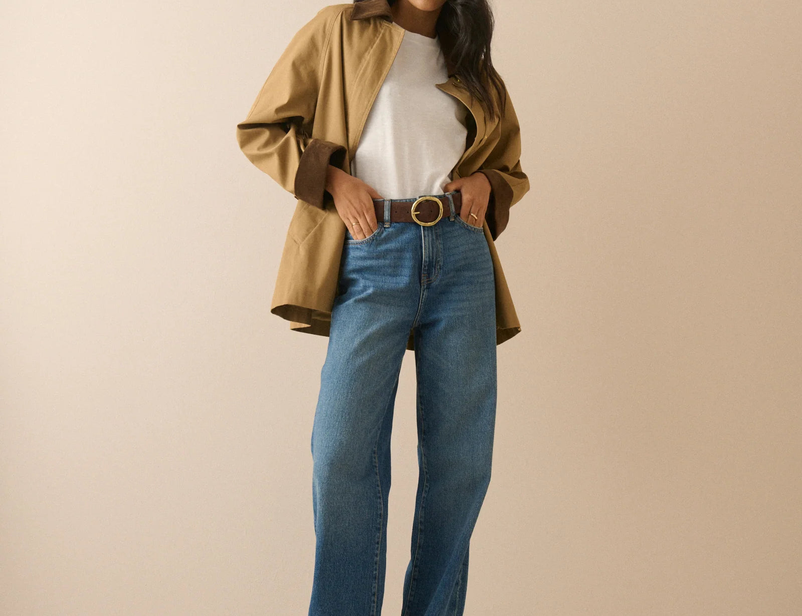 A woman modeling a stylish outfit consisting of a tan overshirt, white t-shirt, blue wide-leg jeans, and a brown belt, standing against a solid beige studio background.