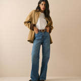 A woman modeling a stylish outfit consisting of a tan overshirt, white t-shirt, blue wide-leg jeans, and a brown belt, standing against a solid beige studio background.