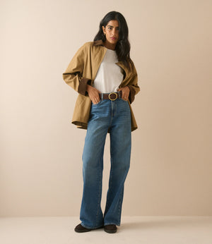 A woman modeling a stylish outfit consisting of a tan overshirt, white t-shirt, blue wide-leg jeans, and a brown belt, standing against a solid beige studio background.