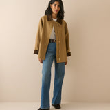 A woman modeling a tan wool-blend jacket with dark brown collar and cuffs, paired with blue jeans and black shoes, standing against a solid beige studio background.