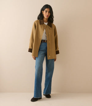 A woman modeling a tan wool-blend jacket with dark brown collar and cuffs, paired with blue jeans and black shoes, standing against a solid beige studio background.