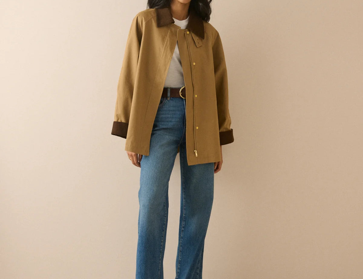 A woman modeling a tan wool-blend jacket with dark brown collar and cuffs, paired with blue jeans and black shoes, standing against a solid beige studio background.