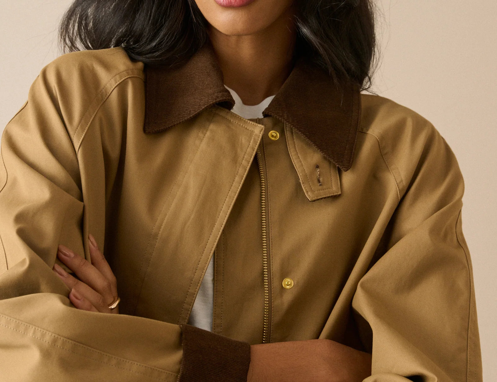 A woman wearing a tan oversized jacket with dark brown collar, cuffs, and zipper trim, paired with a white shirt underneath, set against a solid beige background.