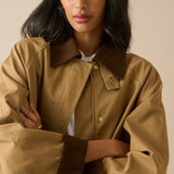 A woman wearing a tan oversized jacket with dark brown collar, cuffs, and zipper trim, paired with a white shirt underneath, set against a solid beige background.