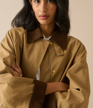 A woman wearing a tan oversized jacket with dark brown collar, cuffs, and zipper trim, paired with a white shirt underneath, set against a solid beige background.