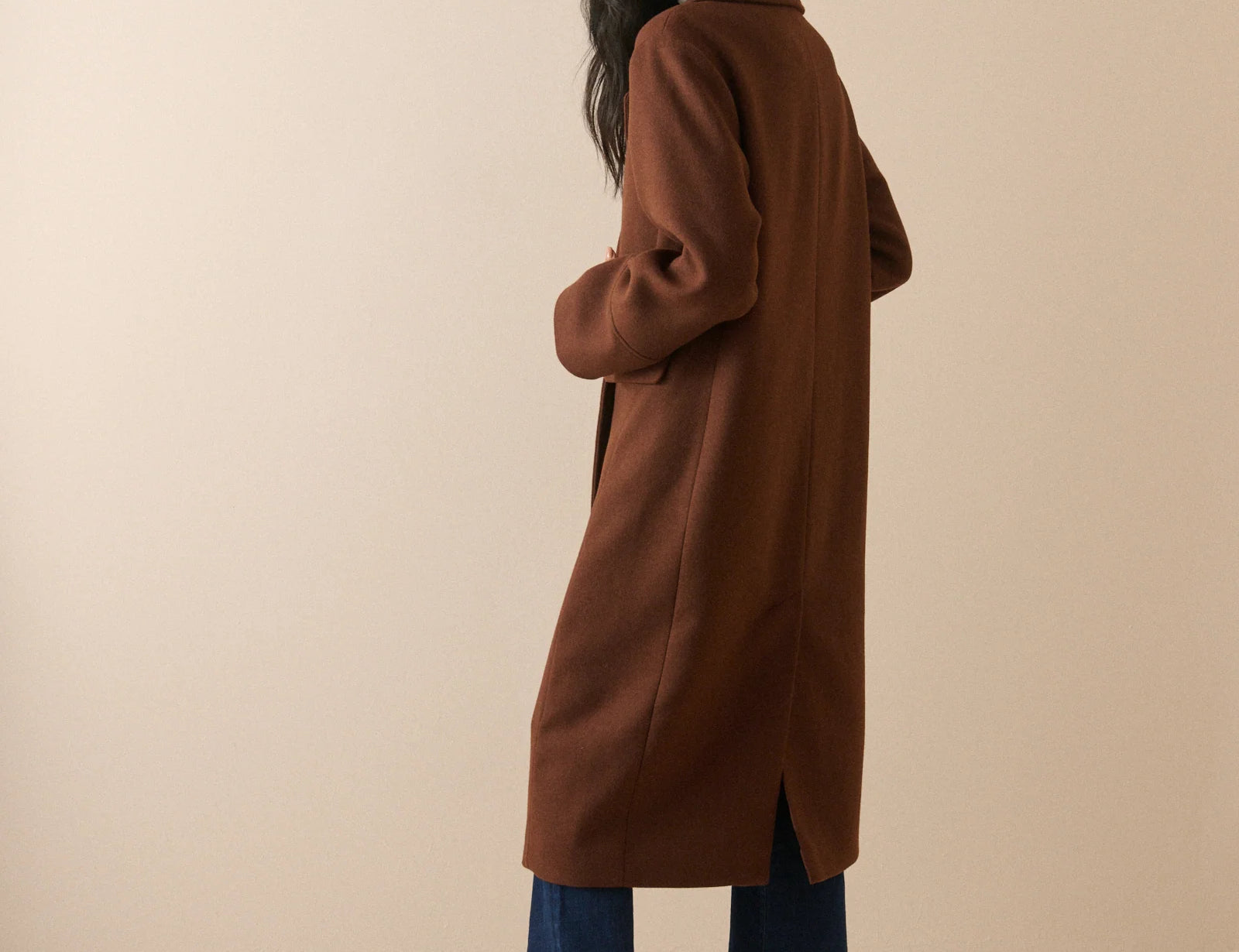 A woman modeling a long brown wool-blend coat, shown from a three-quarters back view. She is wearing blue wide-leg jeans and dark brown heeled boots against a solid beige studio background.