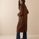 A woman modeling a long brown wool-blend coat, shown from a three-quarters back view. She is wearing blue wide-leg jeans and dark brown heeled boots against a solid beige studio background.