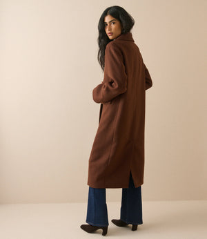 A woman modeling a long brown wool-blend coat, shown from a three-quarters back view. She is wearing blue wide-leg jeans and dark brown heeled boots against a solid beige studio background.