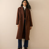 A woman modeling a long brown double-breasted wool coat over a white turtleneck, dark plaid shirt, blue jeans, and black ankle boots, standing against a solid beige studio backdrop.