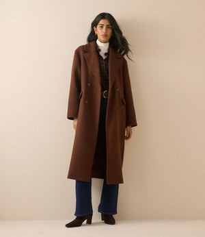 A woman modeling a long brown double-breasted wool coat over a white turtleneck, dark plaid shirt, blue jeans, and black ankle boots, standing against a solid beige studio backdrop.