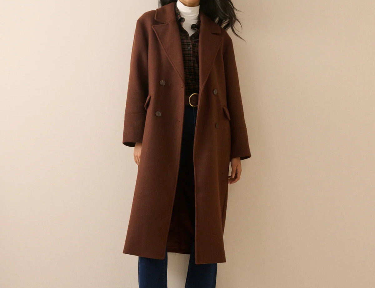 A woman modeling a long brown double-breasted wool coat over a white turtleneck, dark plaid shirt, blue jeans, and black ankle boots, standing against a solid beige studio backdrop.