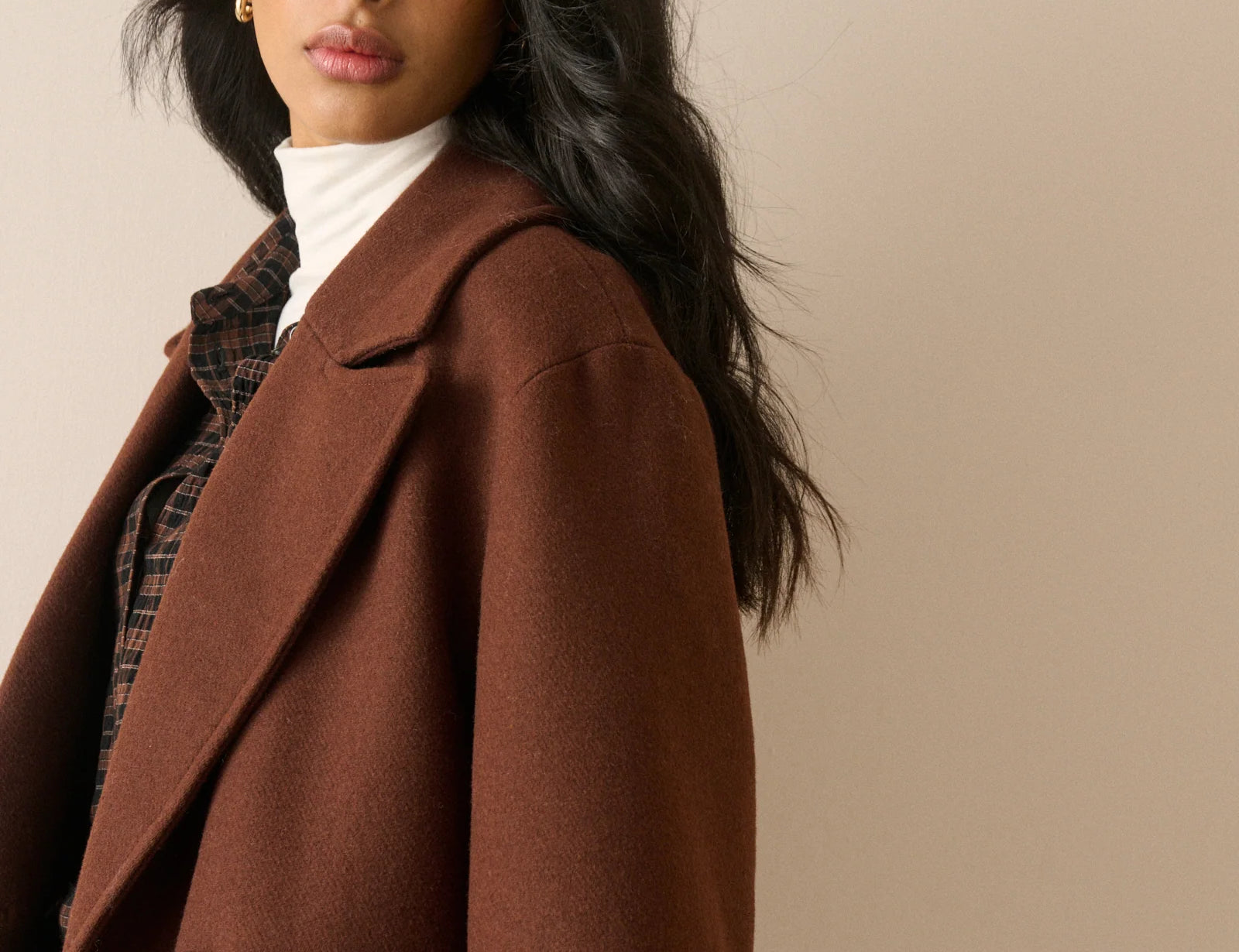 A woman wearing a brown wool-blend blazer over a white turtleneck and plaid shirt, photographed against a plain beige studio background.