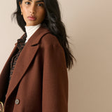 A woman wearing a brown wool-blend blazer over a white turtleneck and plaid shirt, photographed against a plain beige studio background.