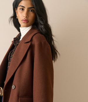A woman wearing a brown wool-blend blazer over a white turtleneck and plaid shirt, photographed against a plain beige studio background.