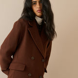 A woman wearing a double-breasted brown wool coat over a cream turtleneck sweater, photographed against a solid beige studio background.
