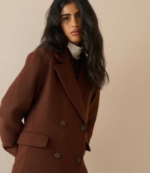 A woman wearing a double-breasted brown wool coat over a cream turtleneck sweater, photographed against a solid beige studio background.