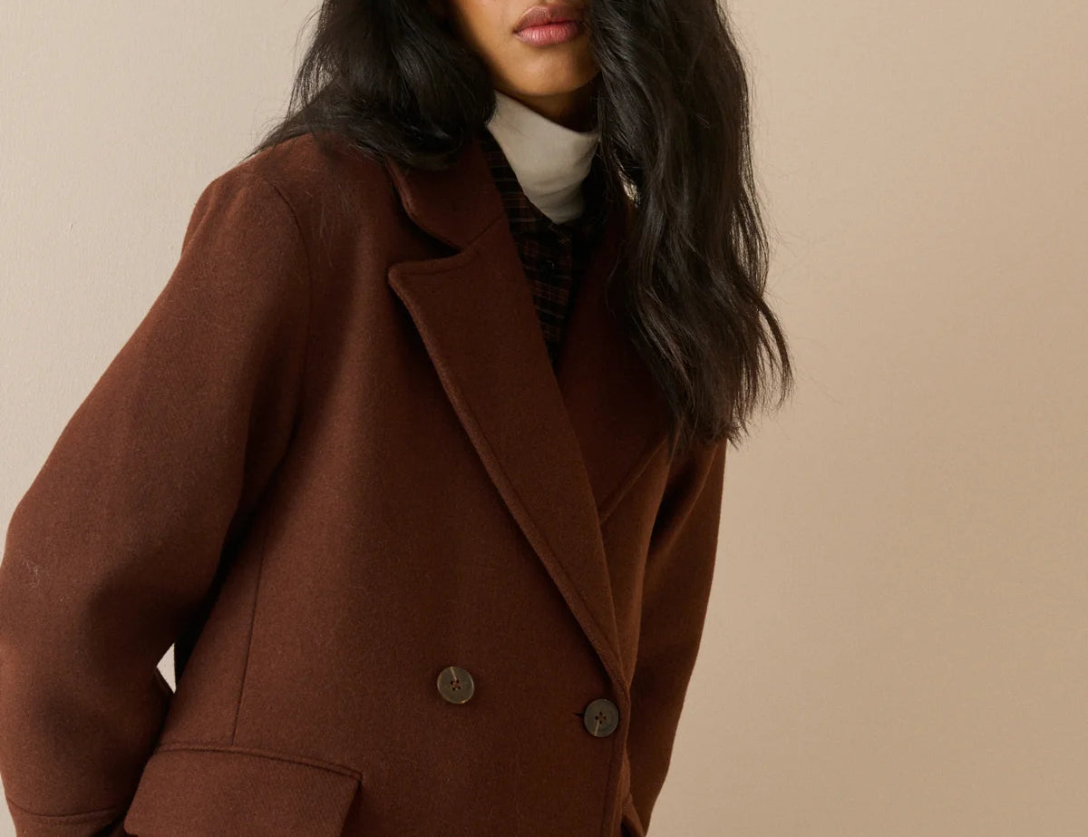 A woman wearing a double-breasted brown wool coat over a cream turtleneck sweater, photographed against a solid beige studio background.