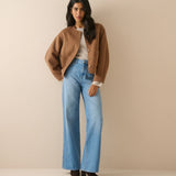 A woman stands against a plain beige studio background wearing a cropped tan jacket, light blue wide-leg jeans, a white top, and black shoes.