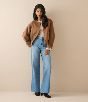 A woman stands against a plain beige studio background wearing a cropped tan jacket, light blue wide-leg jeans, a white top, and black shoes.