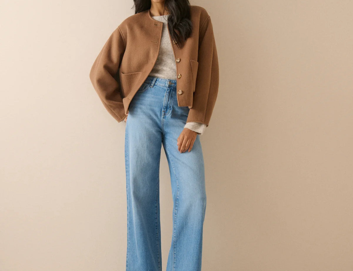 A woman stands against a plain beige studio background wearing a cropped tan jacket, light blue wide-leg jeans, a white top, and black shoes.