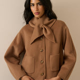 A woman wearing a camel brown wool-blend coat with a large bow detail at the collar, front buttons, and patch pockets, paired with blue jeans, set against a plain beige background.