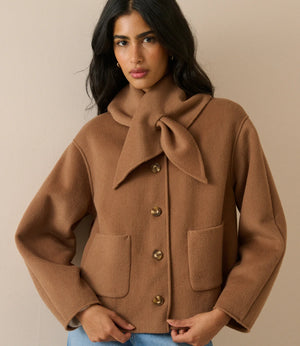 A woman wearing a camel brown wool-blend coat with a large bow detail at the collar, front buttons, and patch pockets, paired with blue jeans, set against a plain beige background.