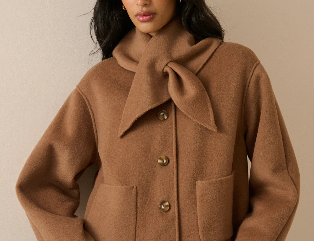 A woman wearing a camel brown wool-blend coat with a large bow detail at the collar, front buttons, and patch pockets, paired with blue jeans, set against a plain beige background.