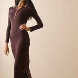 A woman modeling a long-sleeve, bodycon knit midi dress in a deep plum-brown color with contrasting tan collar and placket trim, paired with dark brown boots against a plain beige studio background.