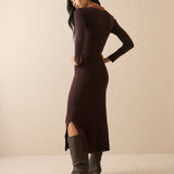 A woman modeling a long-sleeve, bodycon midi dress in deep plum brown, paired with dark gray pointed-toe heeled boots, photographed against a solid beige studio background.