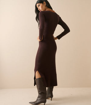 A woman modeling a long-sleeve, bodycon midi dress in deep plum brown, paired with dark gray pointed-toe heeled boots, photographed against a solid beige studio background.