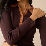 A close-up of a woman wearing a dark brown ribbed long-sleeve henley top with a contrasting tan collar and button placket, set against a solid beige background.