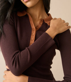 A close-up of a woman wearing a dark brown ribbed long-sleeve henley top with a contrasting tan collar and button placket, set against a solid beige background.