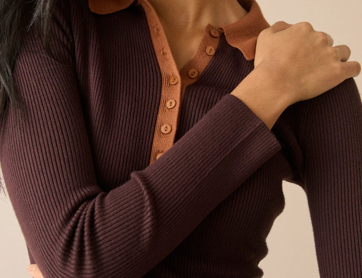 A close-up of a woman wearing a dark brown ribbed long-sleeve henley top with a contrasting tan collar and button placket, set against a solid beige background.