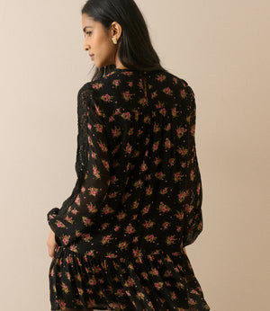 A woman wearing a black long-sleeve mini dress with a pink and yellow floral print, shown from the back against a solid beige background.