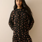 A woman modeling a black long-sleeve mini dress with a dense pink and red floral print, pleated bodice, and ruffled hem, set against a solid beige background.
