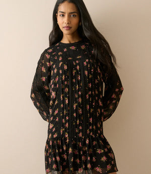 A woman modeling a black long-sleeve mini dress with a dense pink and red floral print, pleated bodice, and ruffled hem, set against a solid beige background.