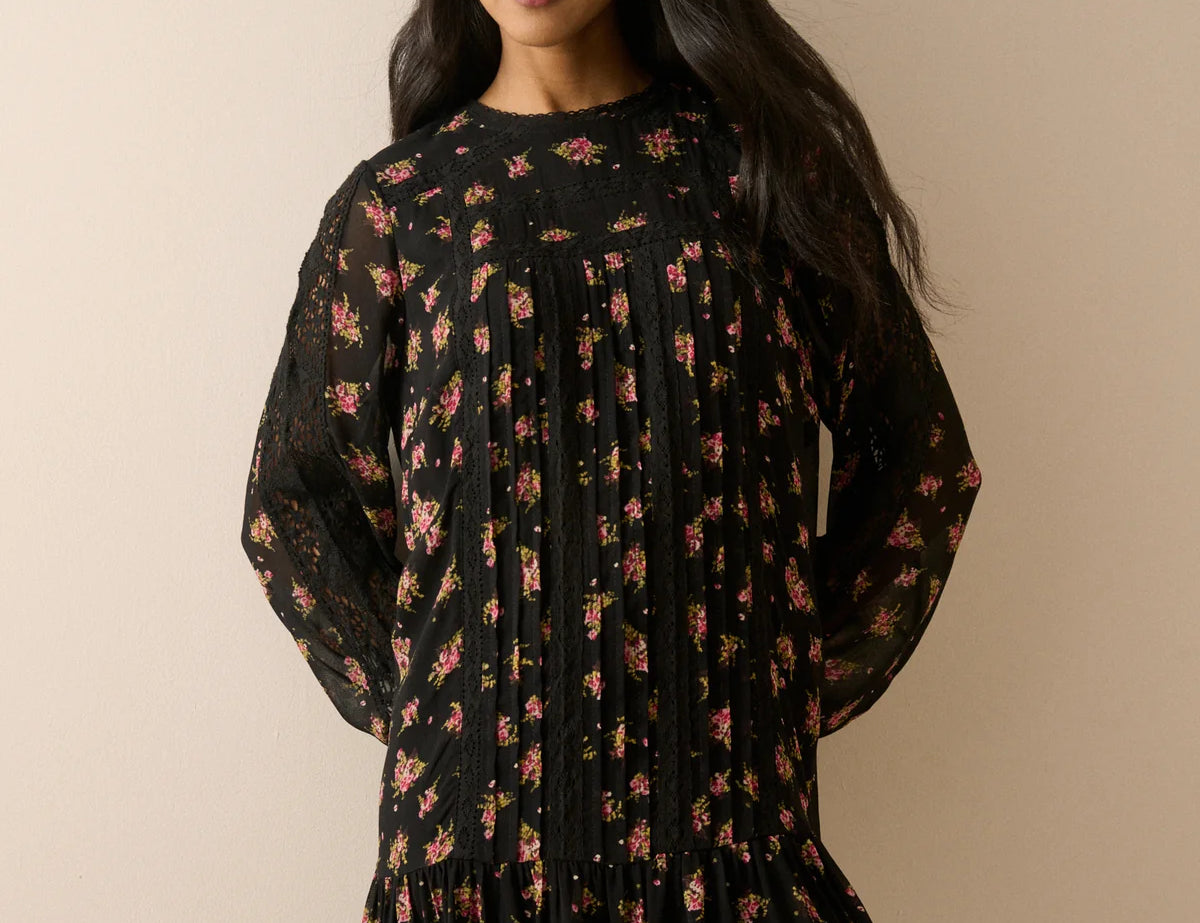 A woman modeling a black long-sleeve mini dress with a dense pink and red floral print, pleated bodice, and ruffled hem, set against a solid beige background.
