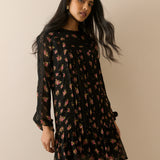 A woman modeling a black long-sleeve mini dress with a pink and red floral pattern, posed against a solid beige background.