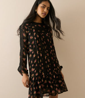 A woman modeling a black long-sleeve mini dress with a pink and red floral pattern, posed against a solid beige background.