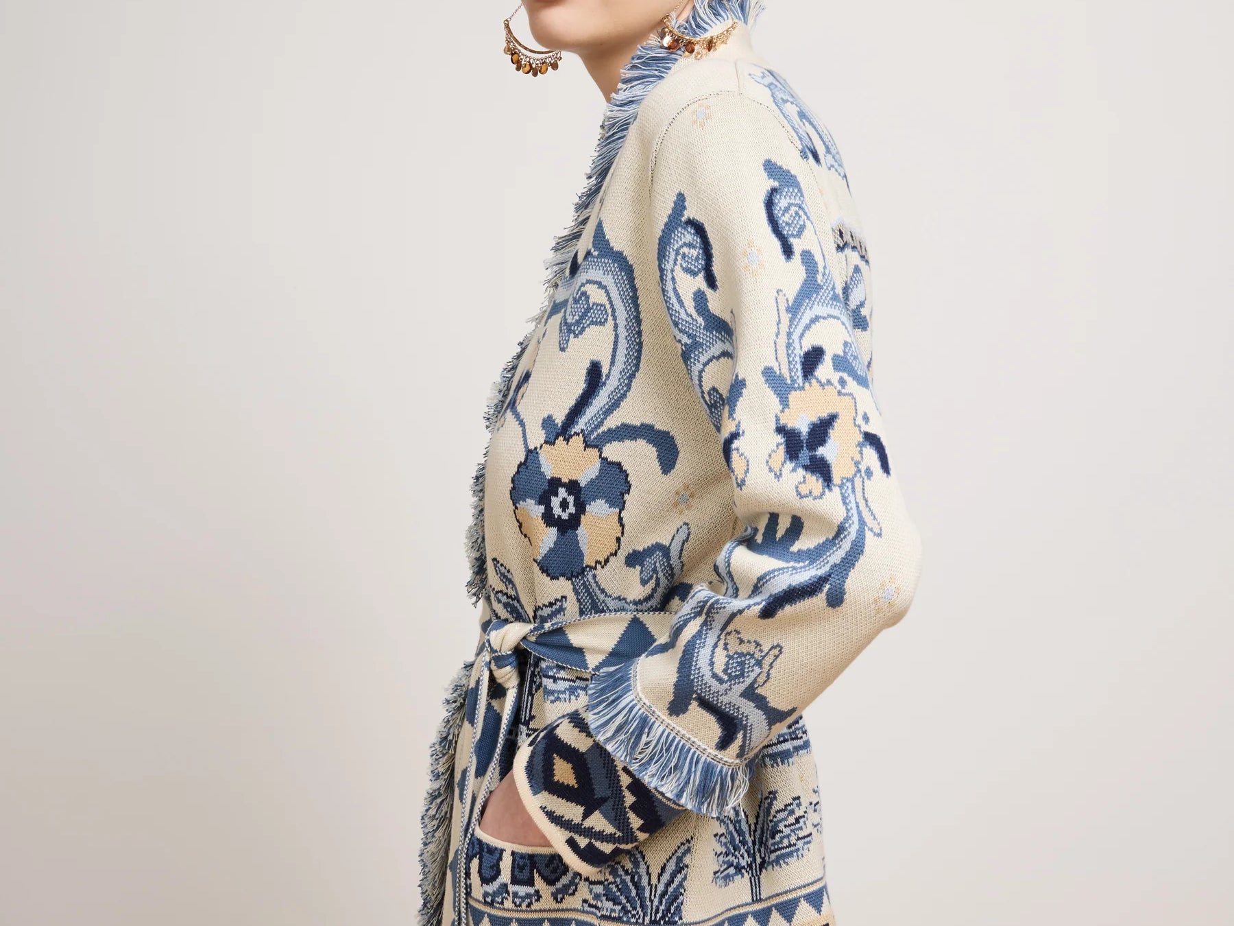 A model wearing a cream-colored knit cardigan with blue and navy patterns, featuring fringe details and a printed design with the words 'MAGNIFICATION LOVE'.
