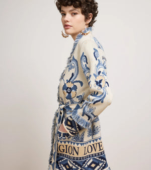 A model wearing a cream-colored knit cardigan with blue and navy patterns, featuring fringe details and a printed design with the words 'MAGNIFICATION LOVE'.