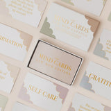 Box of 'The Mind Cards' on a surface with cards displaying text like 'Self Care' and 'Gratitude'.